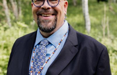 A man in a suit with an indigenour art necklace stands, smiling and looking at the camera, in an outdoor forested setting