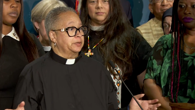 A woman in black clergy vestments stands at a podium with other people of different races and reigious apparel standing in the background