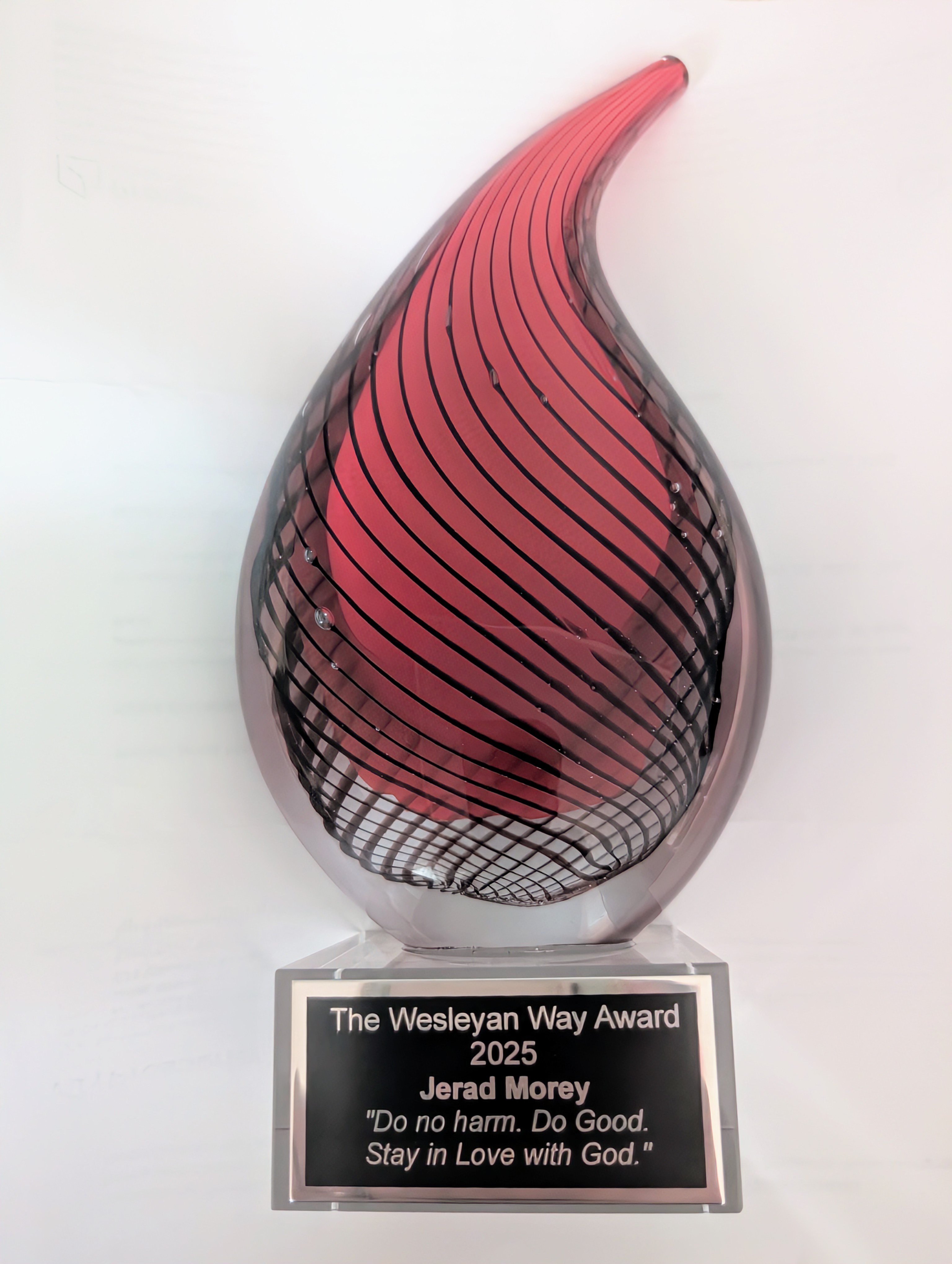 An award in the shape of a flame with a red interior, glass exterior, and a plaque on the bottom reading "The Wesleyan Way Award 2025 Jerad Morey 'Do no harm. Do Good. Stay in Love with God."