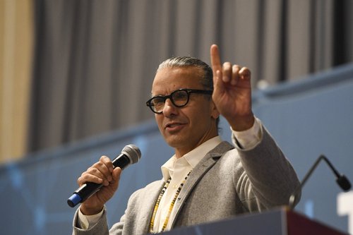 Man with black ponytail and gray blazer stands behind a podium, microphone in one hand and finger raised in another, as though inviting the audience to be patient with a point he's building up to.