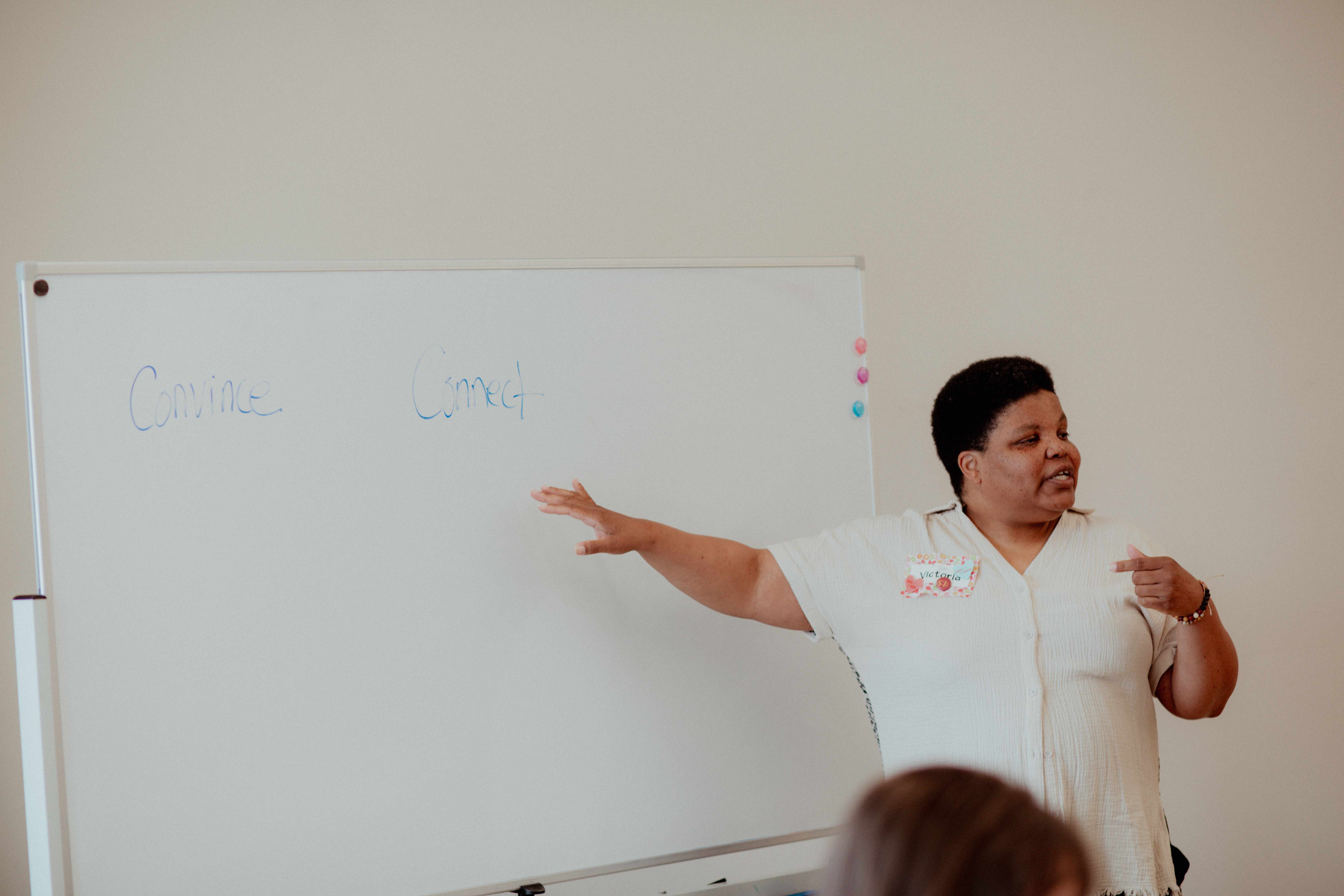 A woman points to a whiteboard while facing an off-screen audience in a brightly-lit room