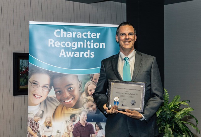 A dashing man holding a plaque stands next to a pull-up graphic stand that reads "Character Recognition Awards"
