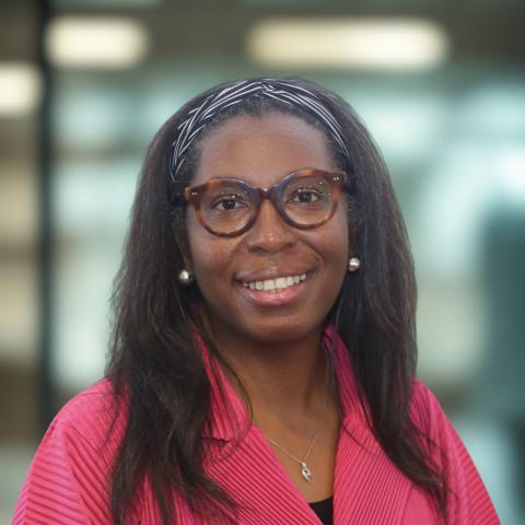 A dark-skinned woman with brown glasses and a black-and-white headband pulling back her past-the shoulders hair wears a pink lined blazer while smiling at the viewer.