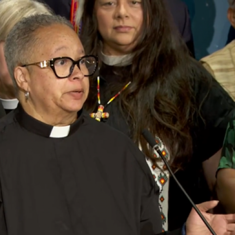 A woman in black clergy vestments stands at a podium with other people of different races and reigious apparel standing in the background