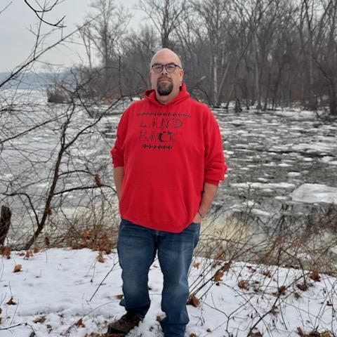 Indigenous man in red #Landback hoodie stands in a wintry, lightly forested landscape