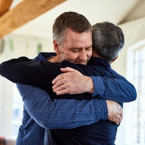 Two middle-aged men hug in a sunlit house.