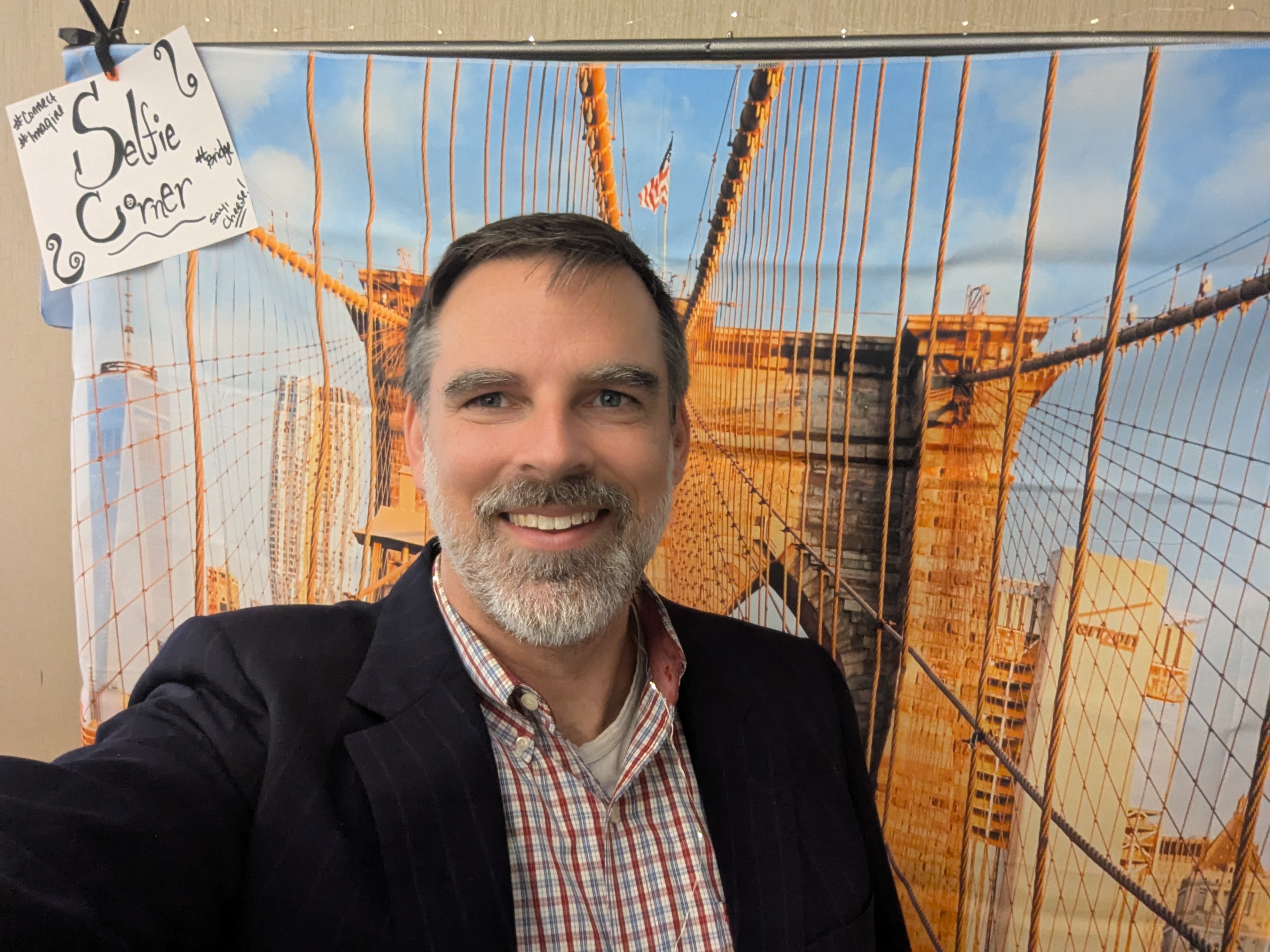 A handsome white man with a beard that does NOT make him look too old stands smiling in front of a large wall hanging of the Brooklyn Bridge.
